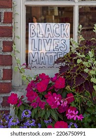 Handwritten Black Lives Matter Sign In Window Pane Of Brick Building, Framed With Flowers Outside Window