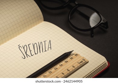 A handwritten inscription "Średnia" on a grille of an open notebook on a wooden countertop, next to a black pencil, lighting of light. (selective focus), translation: mean