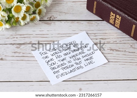 Handwritten note with biblical verse John 3:16 with holy bible book and flowers on wooden table. God's love, forgiveness, salvation, and everlasting life through Jesus Christ, a Christian concept.