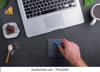 Handwritten notes on black background. 
Hand of a man using laptop computer and chalk.
Top view with copy space.