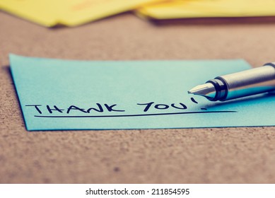 Handwritten Thank You note written on a blue sticky note lying on a cork board with a fountain pen viewed low angle.