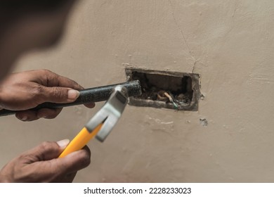 A handyman adjusting a hole in the wall with a chisel and hammer to insert a junction box. Replacing a faulty electrical outlet.