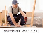 Handyman Assembling Wooden Table With Electric Drill Sitting On Floor During Renovation Indoor. Carpenter Working Making Desk Furnishing House After Renovation. Carpentry Concept