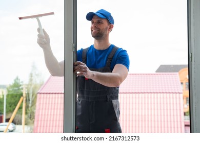 Handyman in blue uniform cleans window glass with squeegee and cleaning supplies in private house outdoors. Cottages view outside new plastic windows