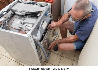 A handyman crouching, using tools and checking components of a broken washing machine, performing maintenance to fix the appliance and restore functionality