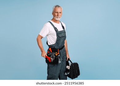 Handyman is holding a wrench and a tool bag while standing in front of a blue background. He is wearing a tool belt and a white t-shirt - Powered by Shutterstock