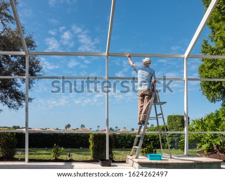 Handyman on ladder cleaning outdoor pool cage enclosure. Screened swimming pool lanai maintenance and screen repair.