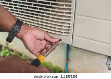 A Handyman Opens Up The Front Hatch Of The Outside Compressor Unit Of Split Type Air Conditioner Unit With A Screwdriver. Repair Or Maintenance Work.