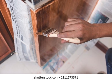 A handyman paints a fresh coat of varnish on the surface of a base kitchen cabinet with a medium sized brush. Home renovation or finishing works. - Shutterstock ID 2111573591