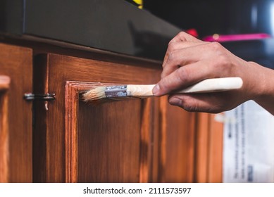 A handyman paints a fresh coat of varnish on the surface of a base kitchen cabinet with a medium sized brush. Home renovation or finishing works.