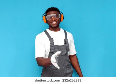 handyman in uniform and construction glasses shows handshake gesture on blue isolated background, african american man in overalls and construction headphones reaches out and accepts