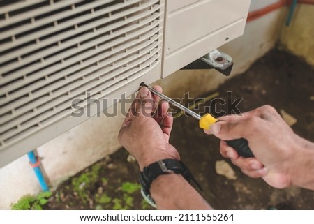 A handyman unscrews the front hatch of the outside compressor unit of split type air conditioner unit. Repair or maintenance work.