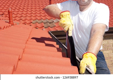 Handyman, Worker Cleaning Gutter On House With Shovel, Roof With Red Tiles And Shingles  As Background.