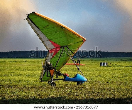 A hang glider with a motor stands on the grass in a field. There are people standing in the distance, a forest and a village are visible. Sunny natural landscape with hangglider.