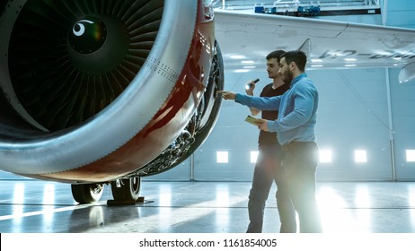 In a Hangar Aircraft Maintenance Engineer Shows Technical Data on Tablet Computer to Airplane Technician, They Diagnose Jet Engine Through Open Hatch. They Stand Near Clean Brand New Plane.