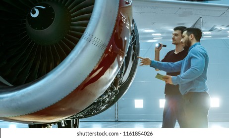 In A Hangar Aircraft Maintenance Engineer Shows Technical Data On Tablet Computer To Airplane Technician, They Diagnose Jet Engine Through Open Hatch. They Stand Near Clean Brand New Plane.