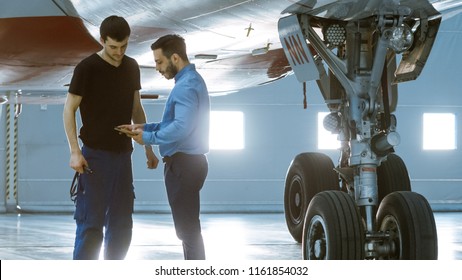 In A Hangar Aircraft Maintenance Engineer Shows  Technical Data On Tablet Computer To Airplane Technician. They Stand Near Clean Brand New Plane.
