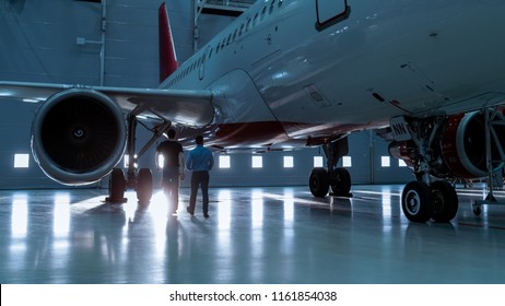 In A Hangar Aircraft Maintenance Engineer Shows  Technical Data On Tablet Computer To Airplane Technician. They Walk Alongside Clean Brand New Plane.