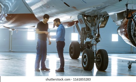 In A Hangar Aircraft Maintenance Engineer Shows  Technical Data On Tablet Computer To Airplane Technician. They Stand Near Clean Brand New Plane.