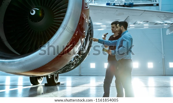 In a Hangar Aircraft Maintenance Engineer Shows
Technical Data on Tablet Computer to Airplane Technician, They
Diagnose Jet Engine Through Open Hatch. They Stand Near Clean Brand
New Plane.