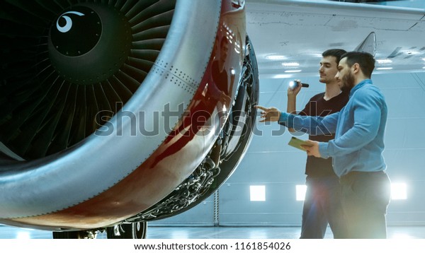 In a Hangar Aircraft Maintenance Engineer Shows\
Technical Data on Tablet Computer to Airplane Technician, They\
Diagnose Jet Engine Through Open Hatch. They Stand Near Clean Brand\
New Plane.