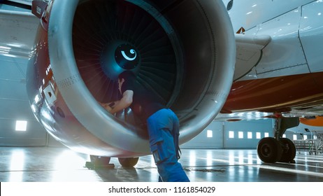 In a Hangar Aircraft Maintenance Engineer/ Technician/ Mechanic Inspects with a Flashlight Airplane's Jet Engine.