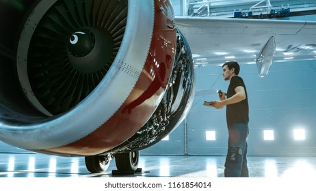 In A Hangar Aircraft Maintenance Young Engineer/ Technician/ Mechanic With Tablet Computer Inspects Airplane Jet Engine. He Opens Engine Hatch And Examines Insides With A Flashlight.