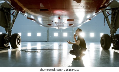 In a Hangar Aircraft Maintenance Young Engineer/ Technician/ Mechanic With Tablet Computer Inspects Airplane Jet Engine. He Opens Engine Hatch and Examines Insides with a Flashlight. - Shutterstock ID 1161854017