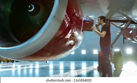 In A Hangar Aircraft Maintenance Young Engineer/ Technician/ Mechanic With Tablet Computer Inspects Airplane Jet Engine. He Opens Engine Hatch And Examines Insides With A Flashlight.