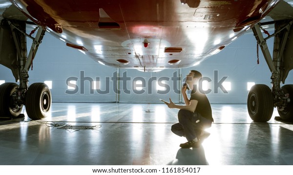 In a Hangar\
Aircraft Maintenance Young Engineer/ Technician/ Mechanic With\
Tablet Computer Inspects Airplane Jet Engine. He Opens Engine Hatch\
and Examines Insides with a\
Flashlight.
