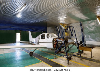 The hangar is a small business auto repair shop. Light single-engine aircraft, in need of engine repair. An aircraft engine is suspended on a chain hoist. - Powered by Shutterstock