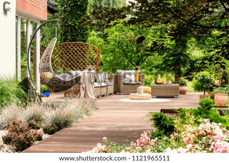 Hanging chair and pouf on wooden veranda in the middle of garden with flowers. Real photo