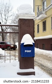 Hanging From A Fence Post Is A Blue Mailbox With A Snow Cap On The Roof. In The Background Is A House And A Car Leaving The Yard.