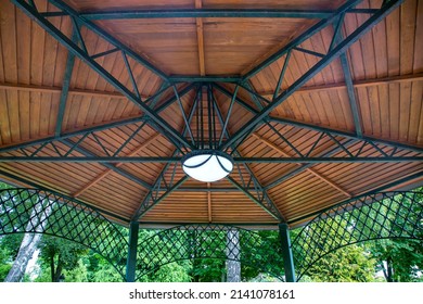 hanging lamp on the ceiling of the gazebo with an iron frame and wooden decorative inserts, a canopy in the backyard with an illuminator among plants close-up.
