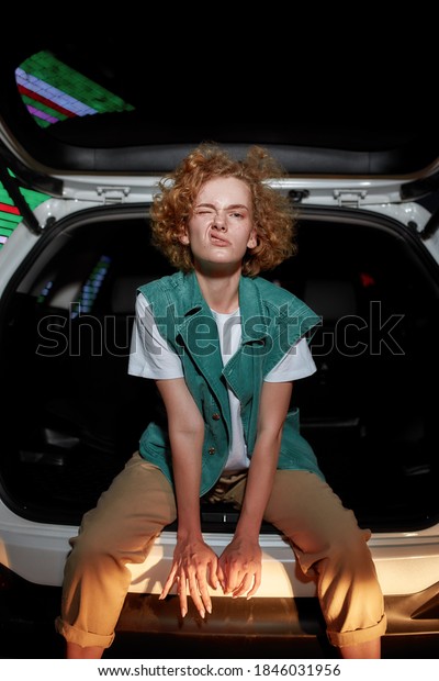 Hanging\
out. An artistic redhead young woman with freckles looking into a\
camera twisting her face and lips while sitting inside of an opened\
car trunk with a led screen behind in the\
evening