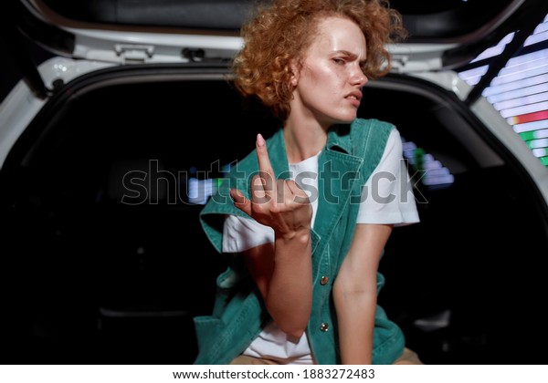Hanging\
out. An artistic redhead young woman with freckles looking into a\
camera with her middle finger out while sitting inside of an opened\
car trunk with a led screen behind in the\
evening