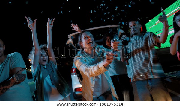 Hanging out. A casually dressed young white\
long haired guy with his firends celebrate a night with opening a\
confetti cracker outside on a parking site with a big led screen on\
a background