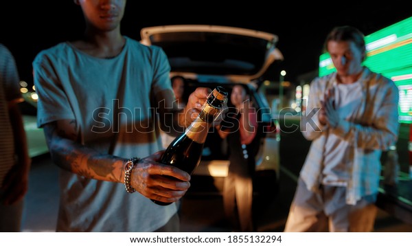 Hanging out. A close up of a young\
tattooed guy opening a bottle of champagne in a group of friends\
with a car and a big led screen on a background at\
night