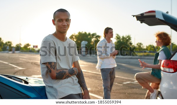 Hanging out. A dark-skinned tattooed man smiling\
looking into a camera standing outside leaning against a car on a\
parking site with his friends on a background speaking to each\
other on a meeting