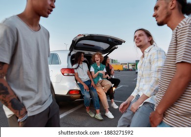 Hanging Out. A Group Of Three Young Well-dressed Ladies Of Different Nationalities Sitting In An Opened Trunk Outside On A Parking Site Smiling And Laughing With Guys On A Front Line During A Sunset