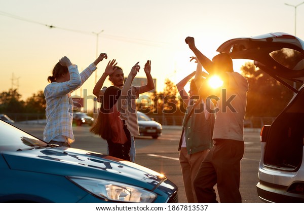 Hanging out. A group of young\
commonly dressed friends having a party dancing to music together\
outside on a parking site near their cars during a beautiful\
sunrise