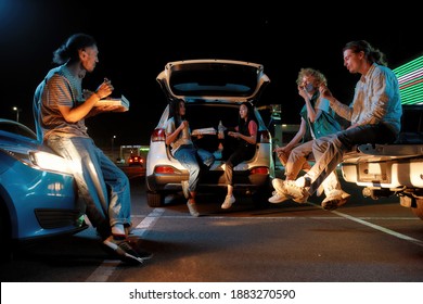 Hanging out. A group of young fashionably dressed friends of different nationalities sitting in opened car trunks in front of each other outside on a parking site eating pizza at night