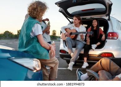 Hanging out. A group of young well-dressed friends of different nationalities sitting on car trunks in front of each other on a parking site drinking beer eating chips playing guitar during a sunset - Powered by Shutterstock