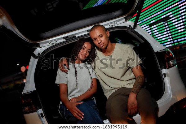 Hanging out. Two young well-dressed dark-skinned\
friends of different genders sitting in an opened car trunk looking\
into a camera hugging each other with a led screen behind in the\
evening