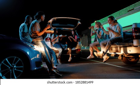 Hanging Out. Young Casually Dressedfriends Of Different Nationalities Sitting In Opened Car Trunks In Front Of Each Other Outside On A Parking Site Happy To Eat Pizza And To Drink Beer At Night