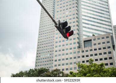 Hanging Red Lighted Traffic Light On The Background Of The Green Trees Tops And A Large Skyscraper In Singapore. View From Below. Horizontal.