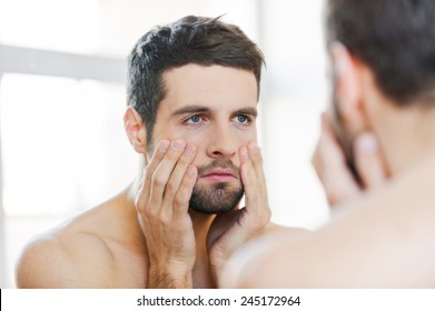 Hangover After Night Out. Frustrated Young Shirtless Man Touching His Face And Looking At Himself While Standing Against A Mirror 