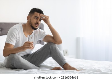 Hangover Concept. Portrait Of Sick Young Arab Guy Holding Glass Of Water And Touching Head While Sitting In Bed At Home, Upset Middle Eastern Man Feeling Unwell, Having Headache, Copy Space