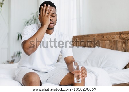Hangover Concept. Sleepy Black Guy Touching Aching Head Holding Glass Of Water Sitting In Bed At Home In The Morning