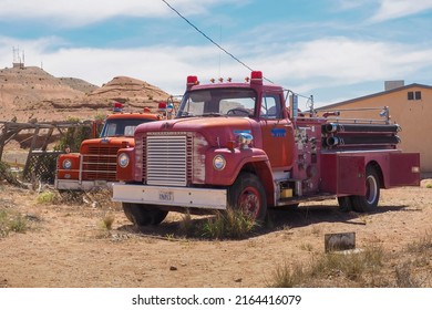 Hanksville, USA - 06.05.2022: Red Vintage Fire Trucks Parked By An Old Building In The Utah Desert. Sandy Hills In The Background. Hot Sunny Day Of Spring. Veteran Fire Fighter Vehicle.